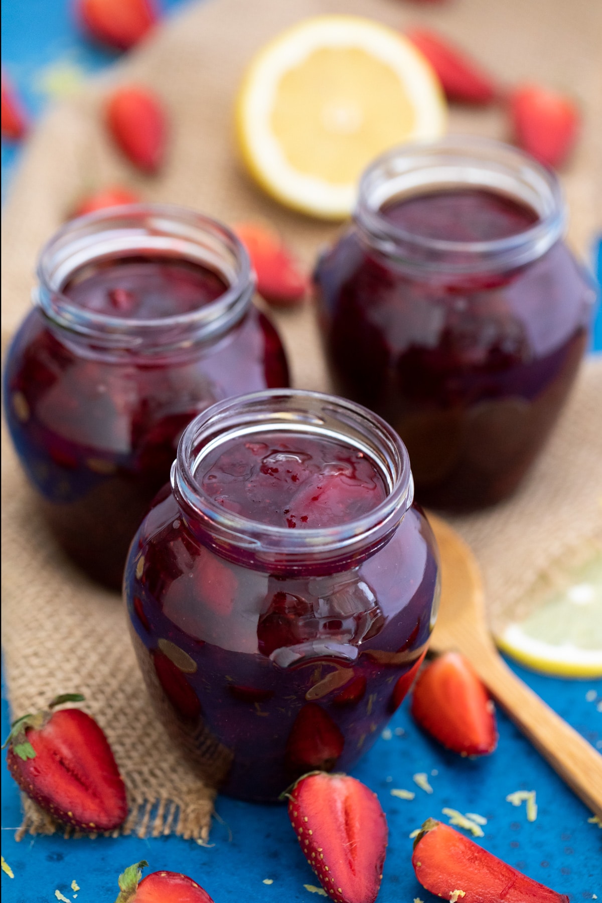 Three jars of jam sitting on blue and burlap napkin with sliced berries on table