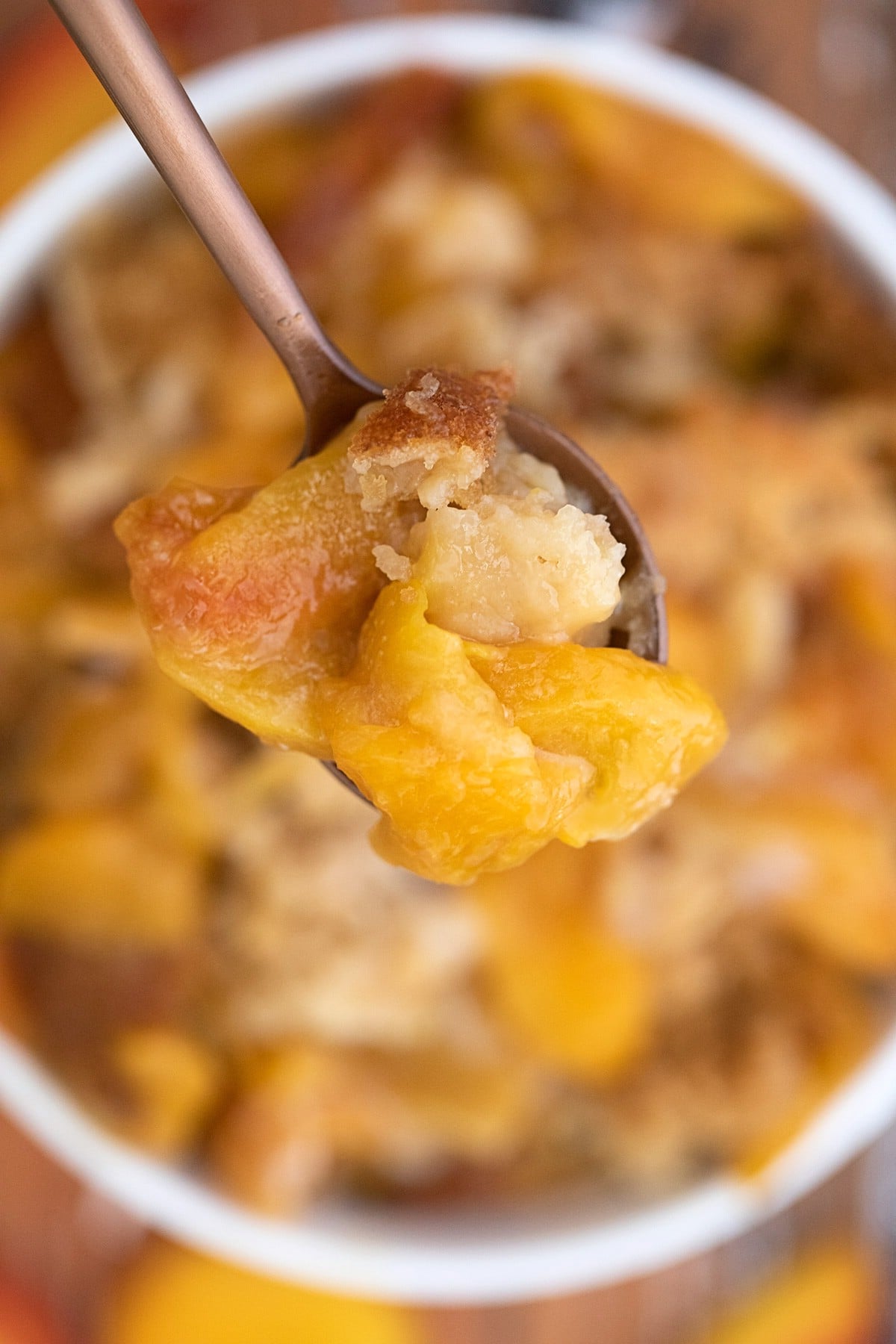 Spoon of peach cobbler hovering over large white bowl of cobbler