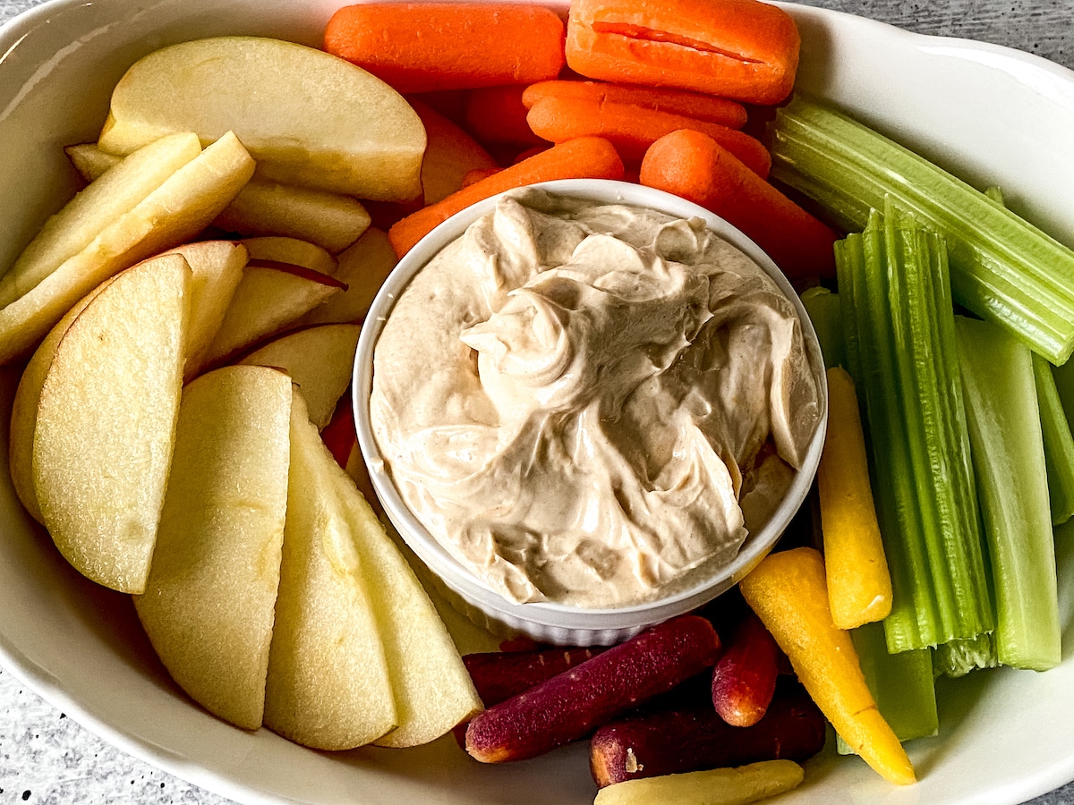 White oval platter with apples carrots and celery alongside bowl of nut butter dip