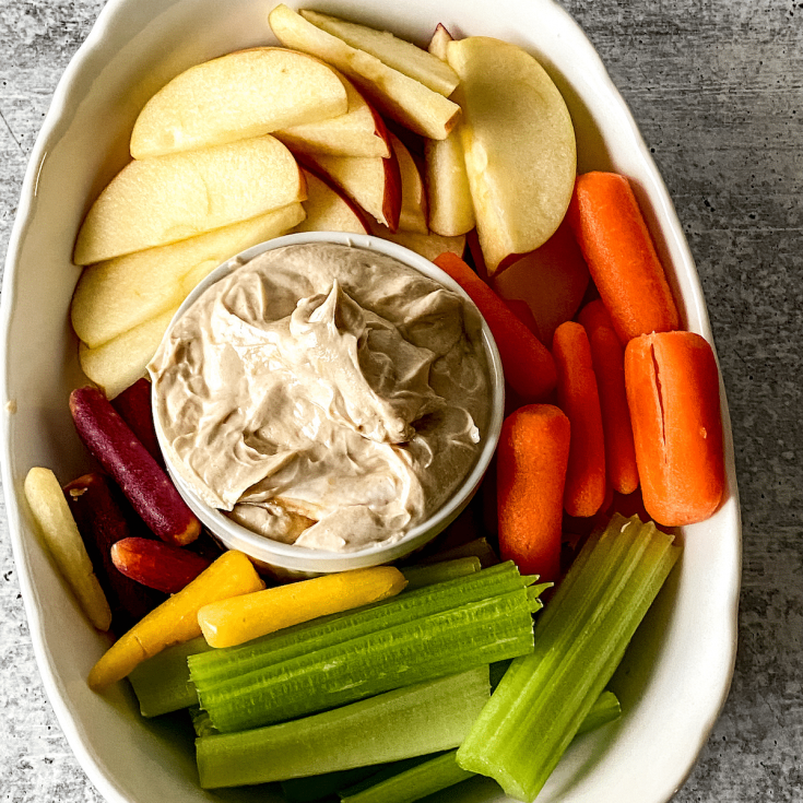 White bowl with vegetables and fruit surrounding a glass bowl of nutbutter dip