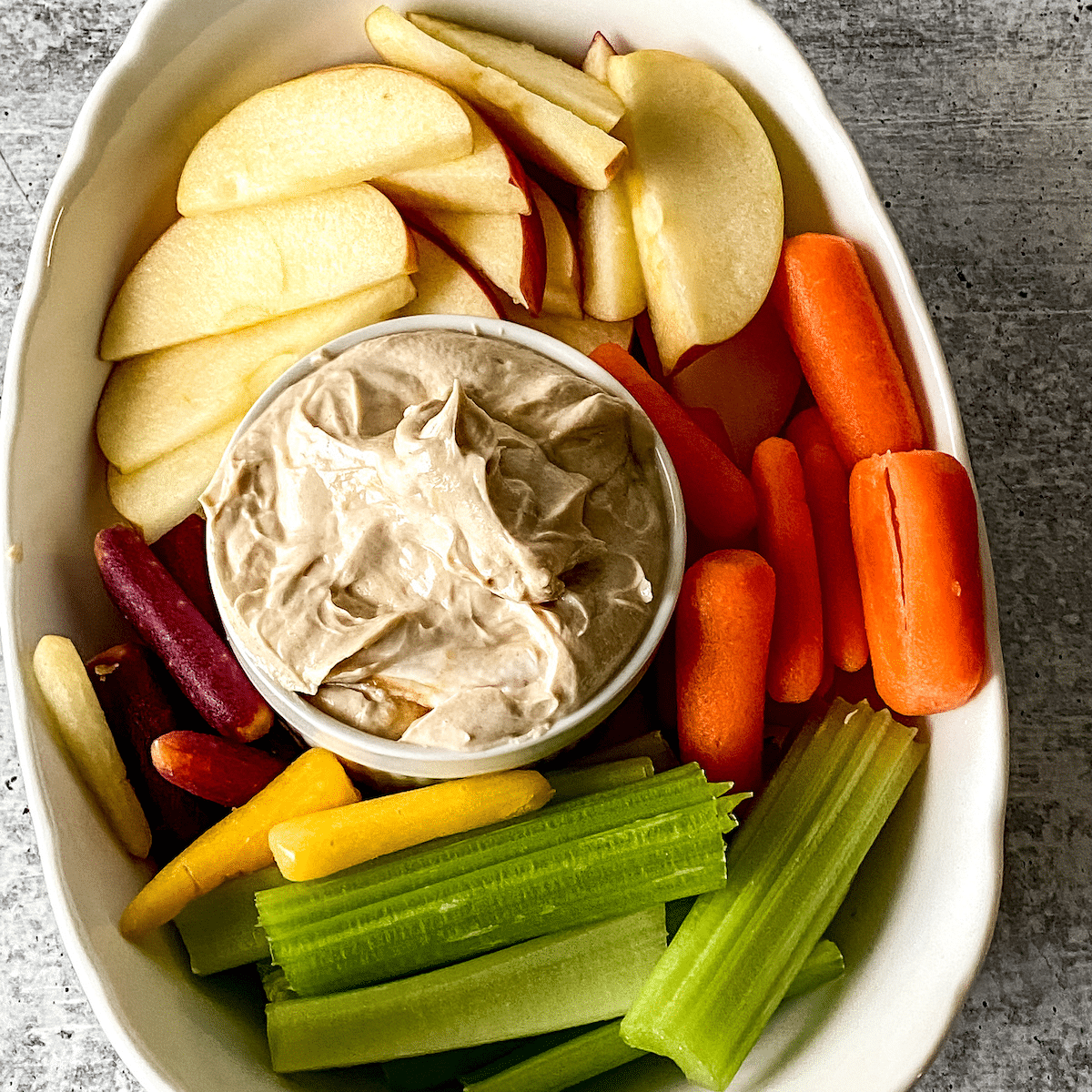 White bowl with vegetables and fruit surrounding a glass bowl of nutbutter dip