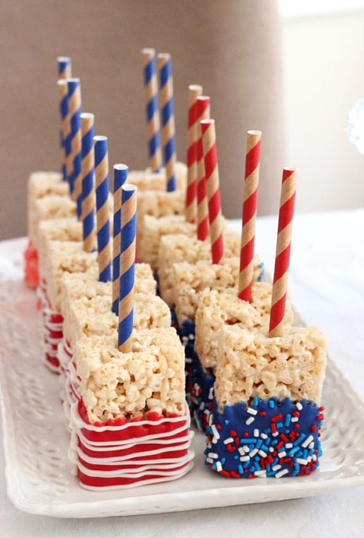 Squares of rice krispie treats dipped in red and blue chocolate with sprinkles and paper straws on top