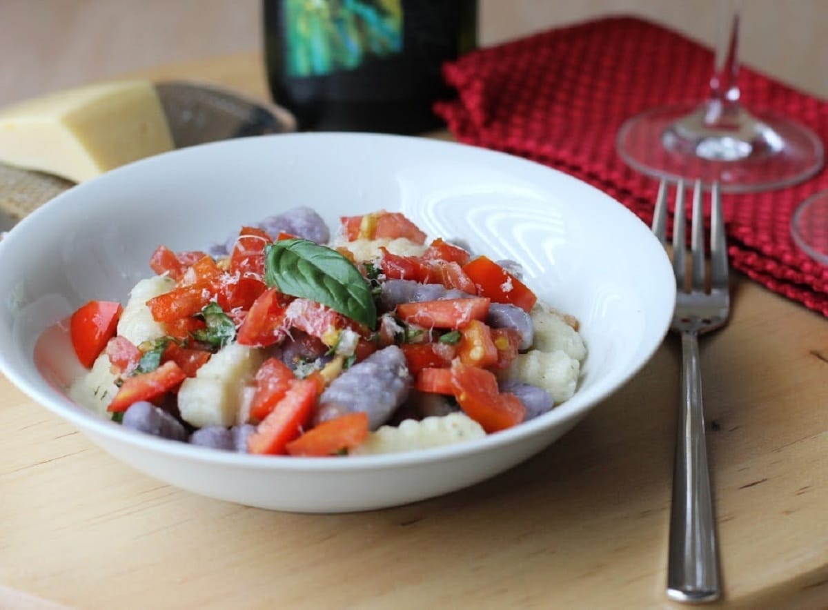 Shallow white bowl of purple gnocchi with red tomatoes next to red napkin and fork