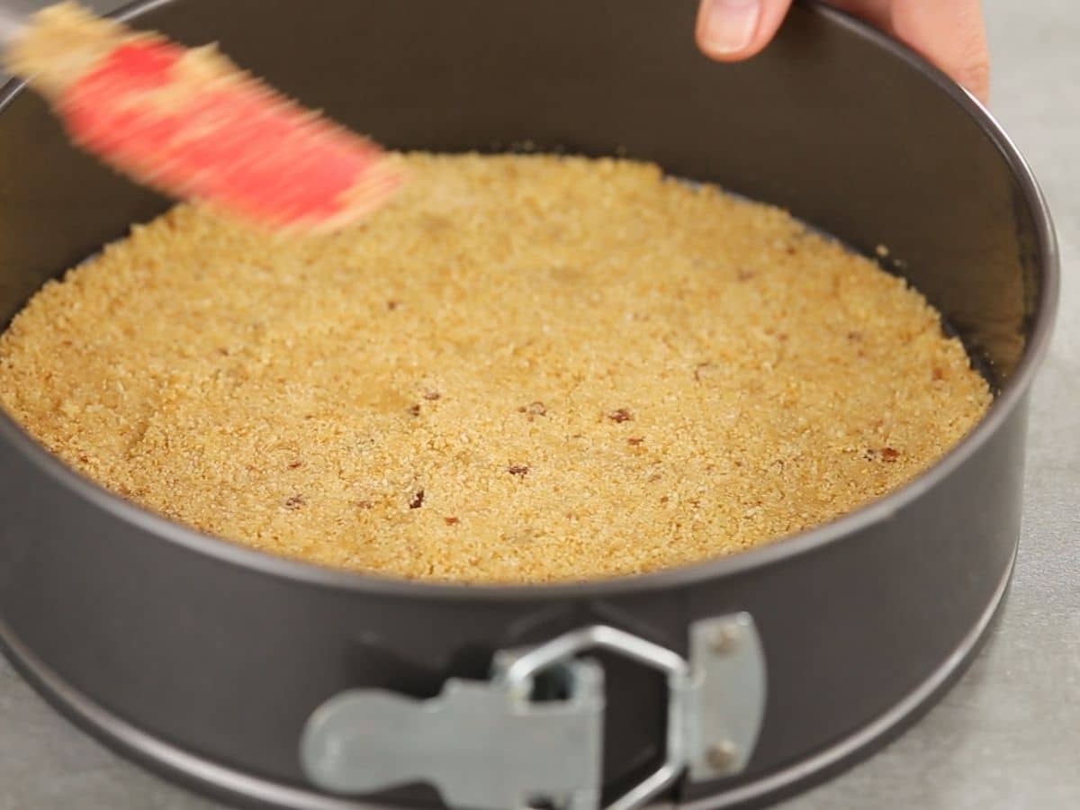 Red spatula pressing graham cracker crust into springform pan