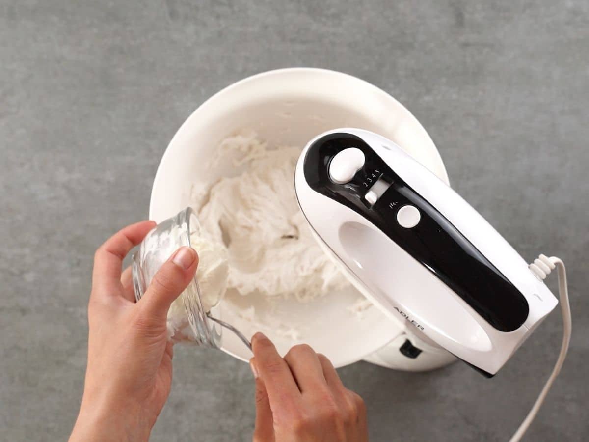 Hand pouring sour cream into white bowl with mixer attached