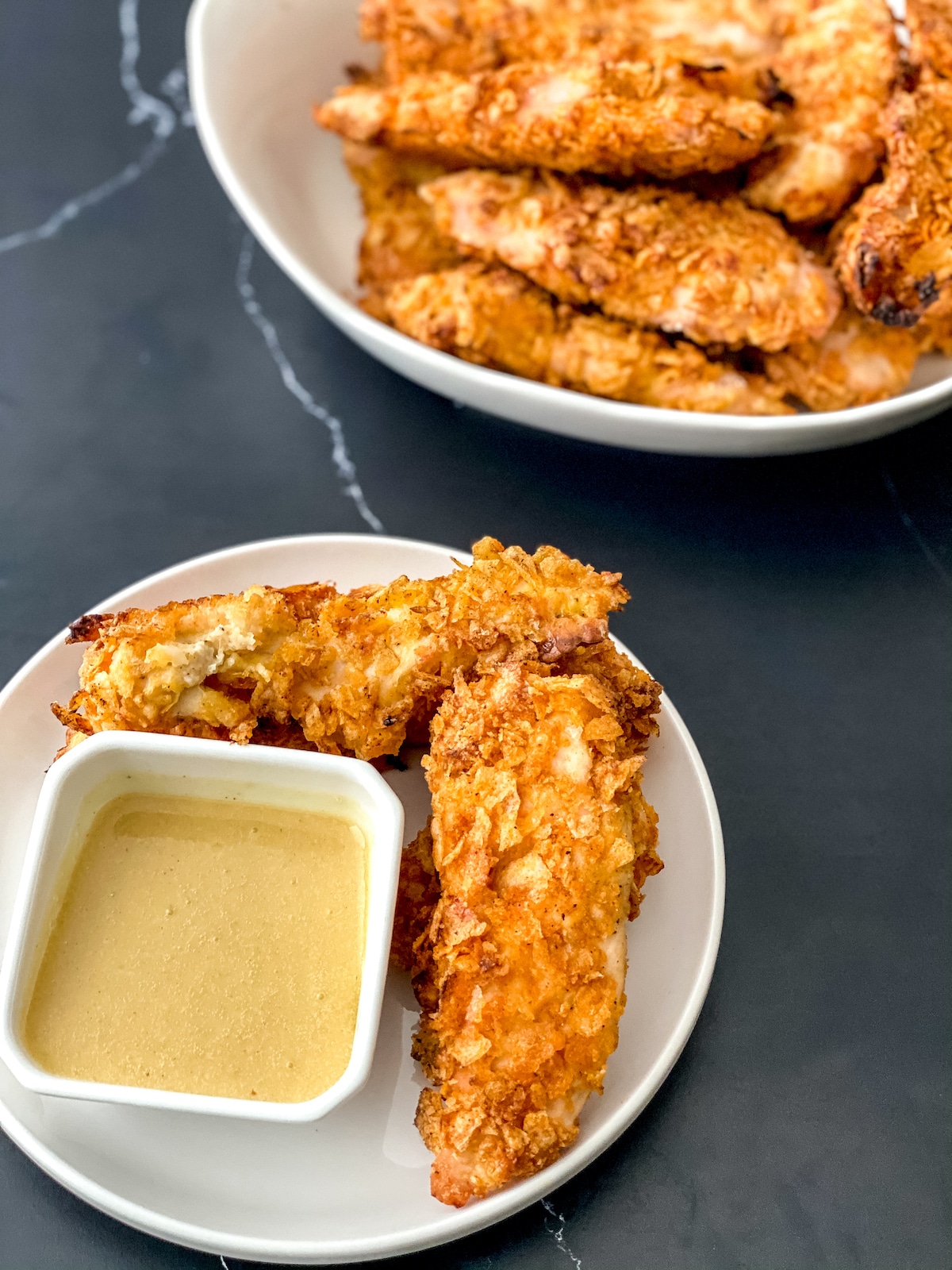 Overhead image of white plate of chicken tenders with bowl of sauce on black table