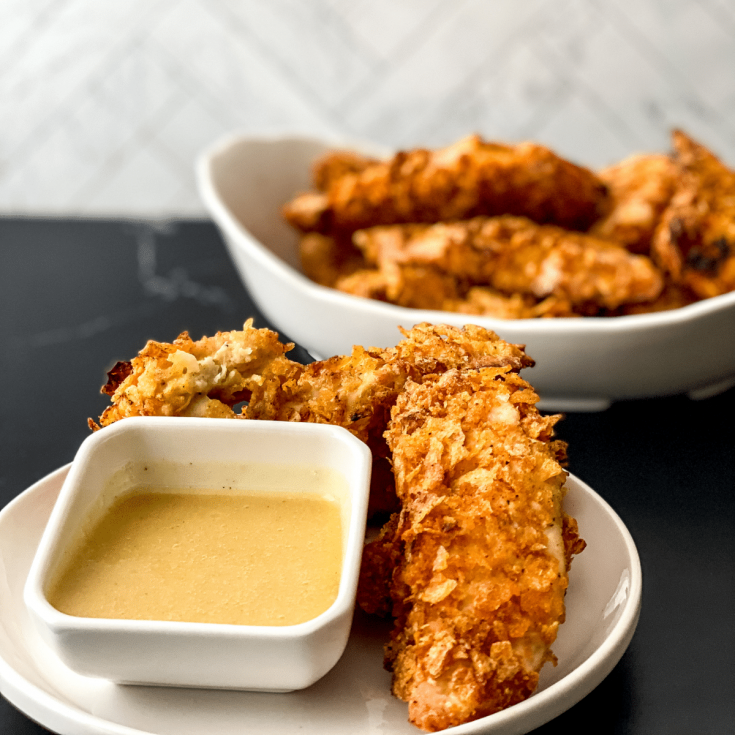 White bowl of chicken tenders on black surface with white saucer of tenders including small bowl of honey mustard