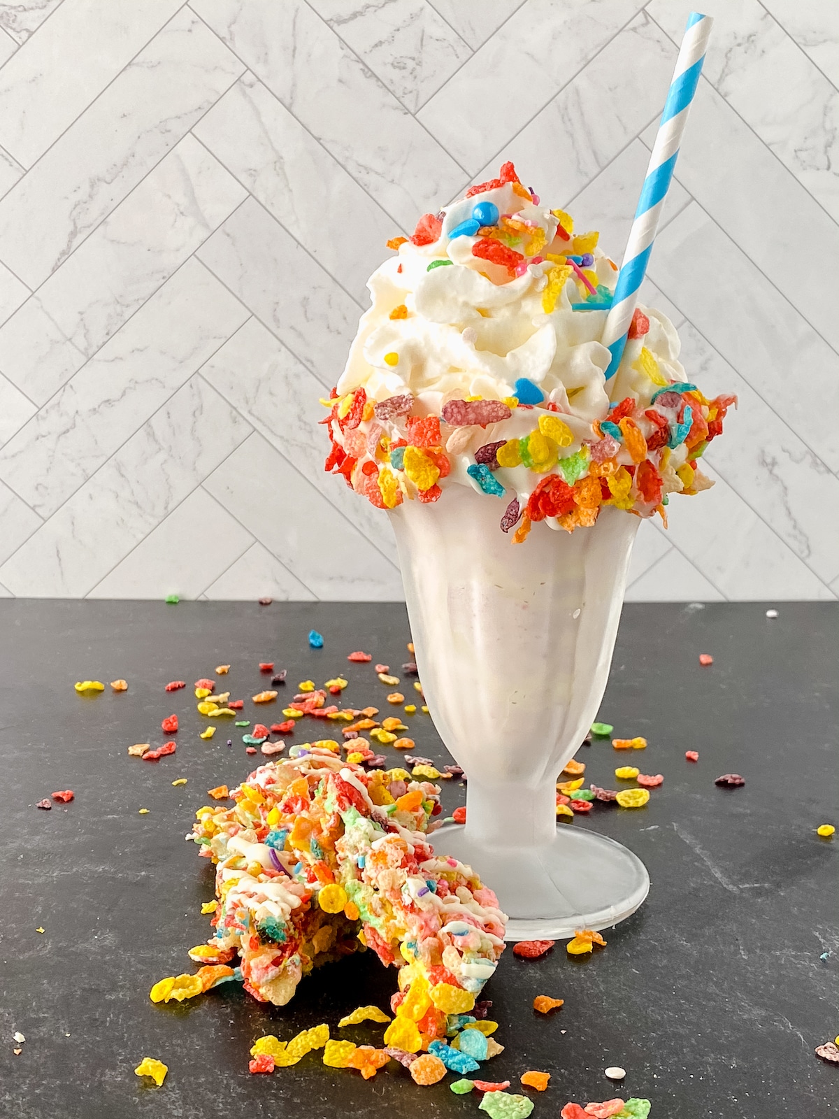 Tall soda glass filled with white milkshake on black table with colorful cereal on top of glass