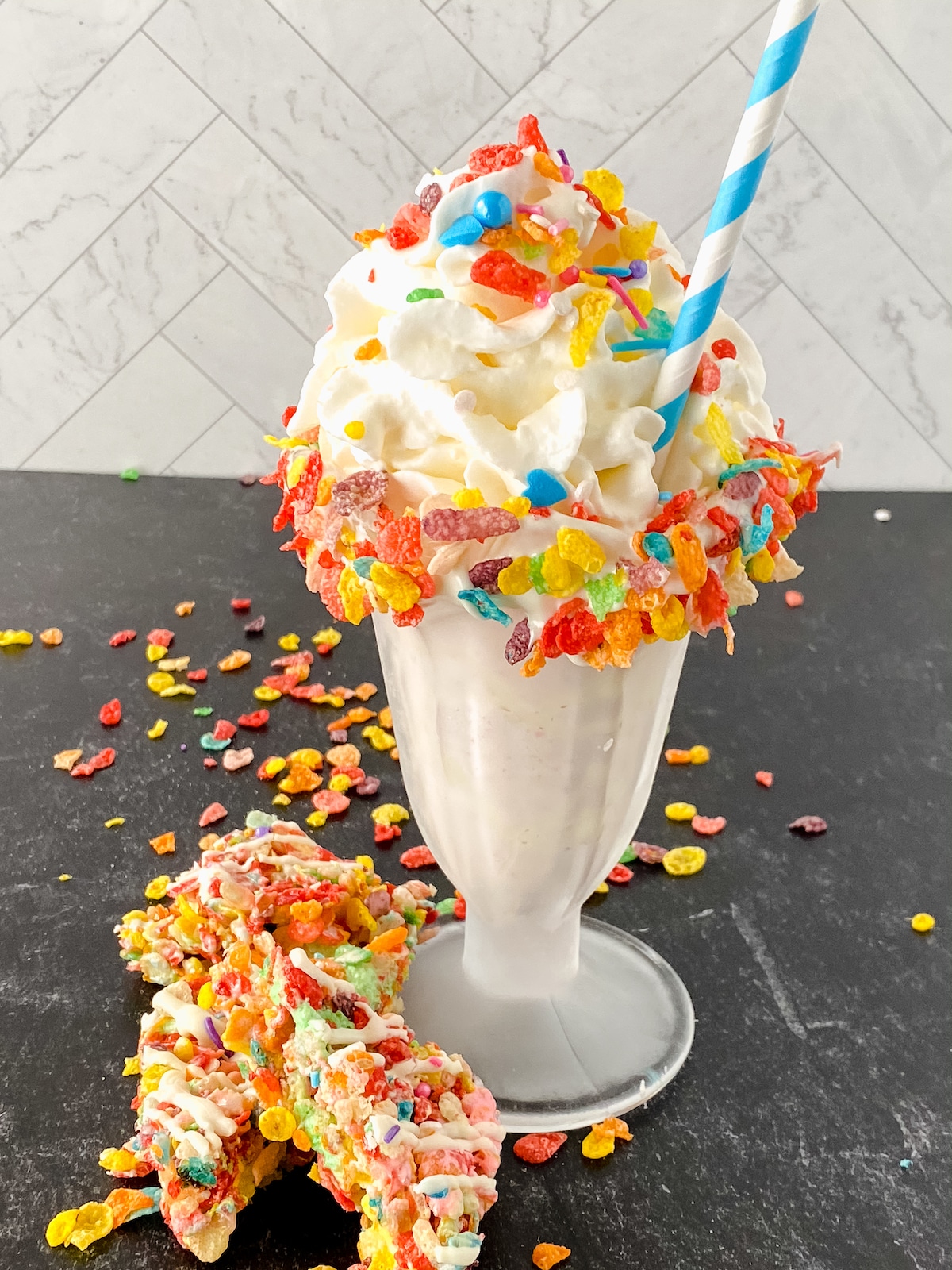 Milkshake in tall glass topped with whipped cream and fruity pebbles on table with additional cream and cereal on side