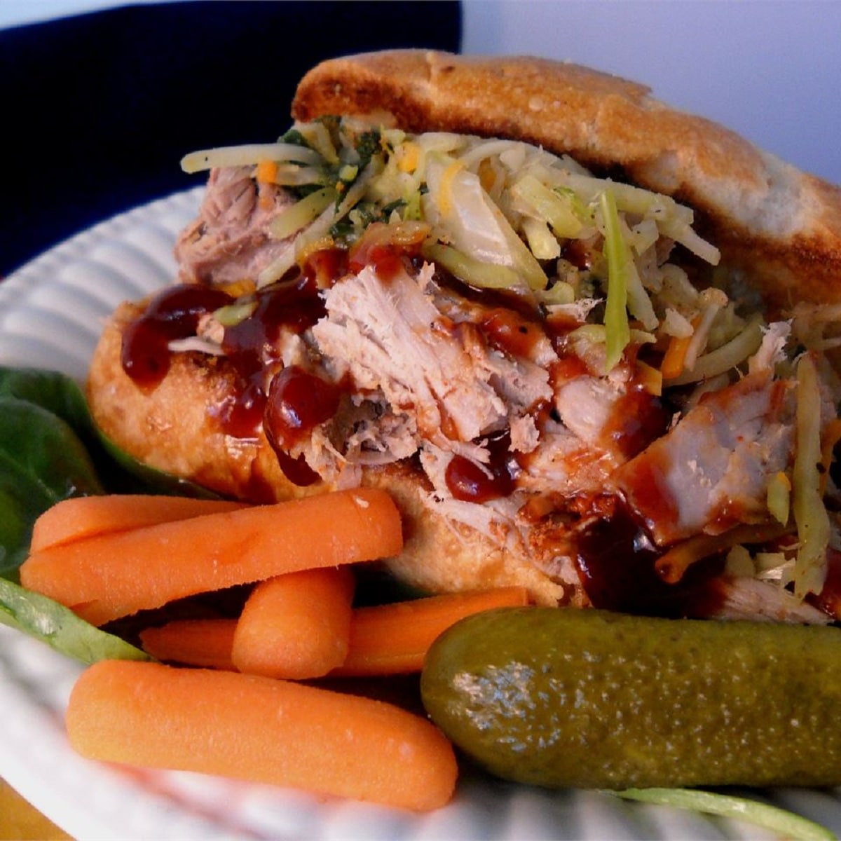 Big sandwich with shredded meat and sauce on white plate next to carrots and pickles