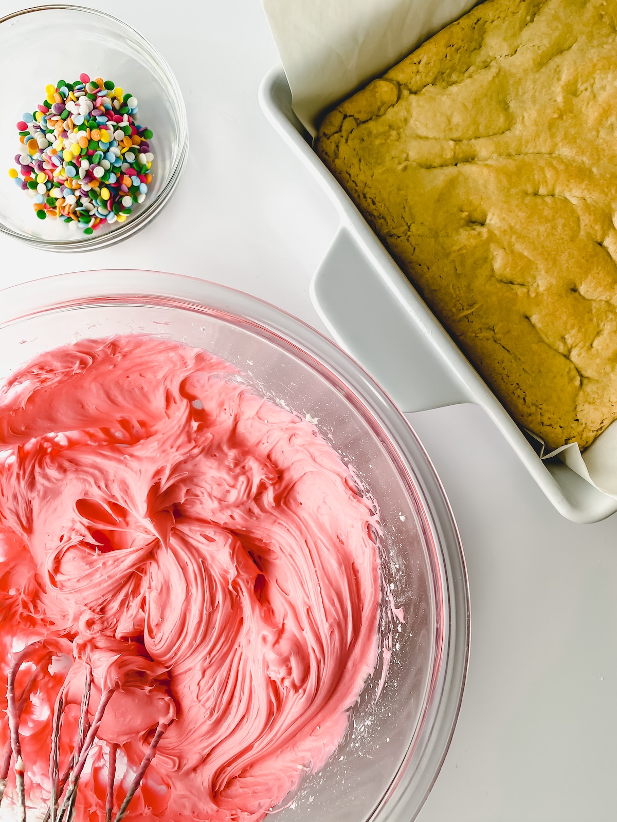 Bowl of pink icing with cake pan beside it and bowl of sprinkles