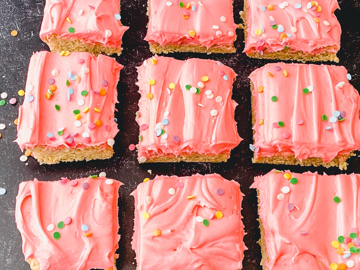 Pan of cookie bars sliced and on black table topped with pink frosting