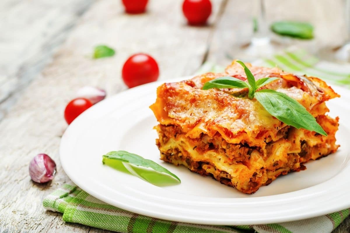 A serving of lasagna on a white plate with cherry tomatoes on the background