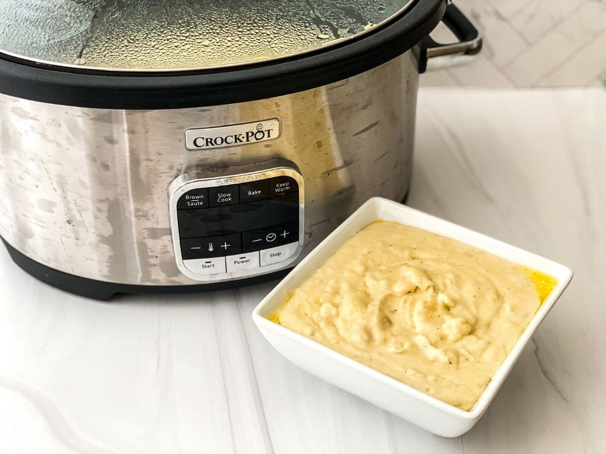 Silver slow cooker on white counter by square bowl of potatoes
