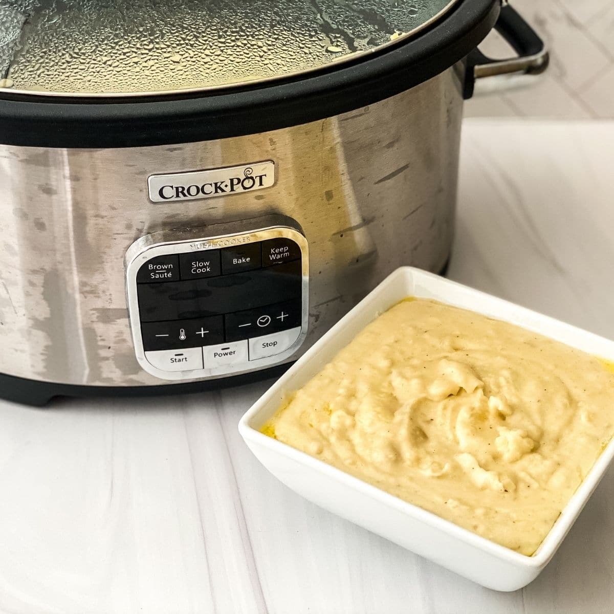 Silver crock Pot on white counter with white square bowl of creamy potatoes in front