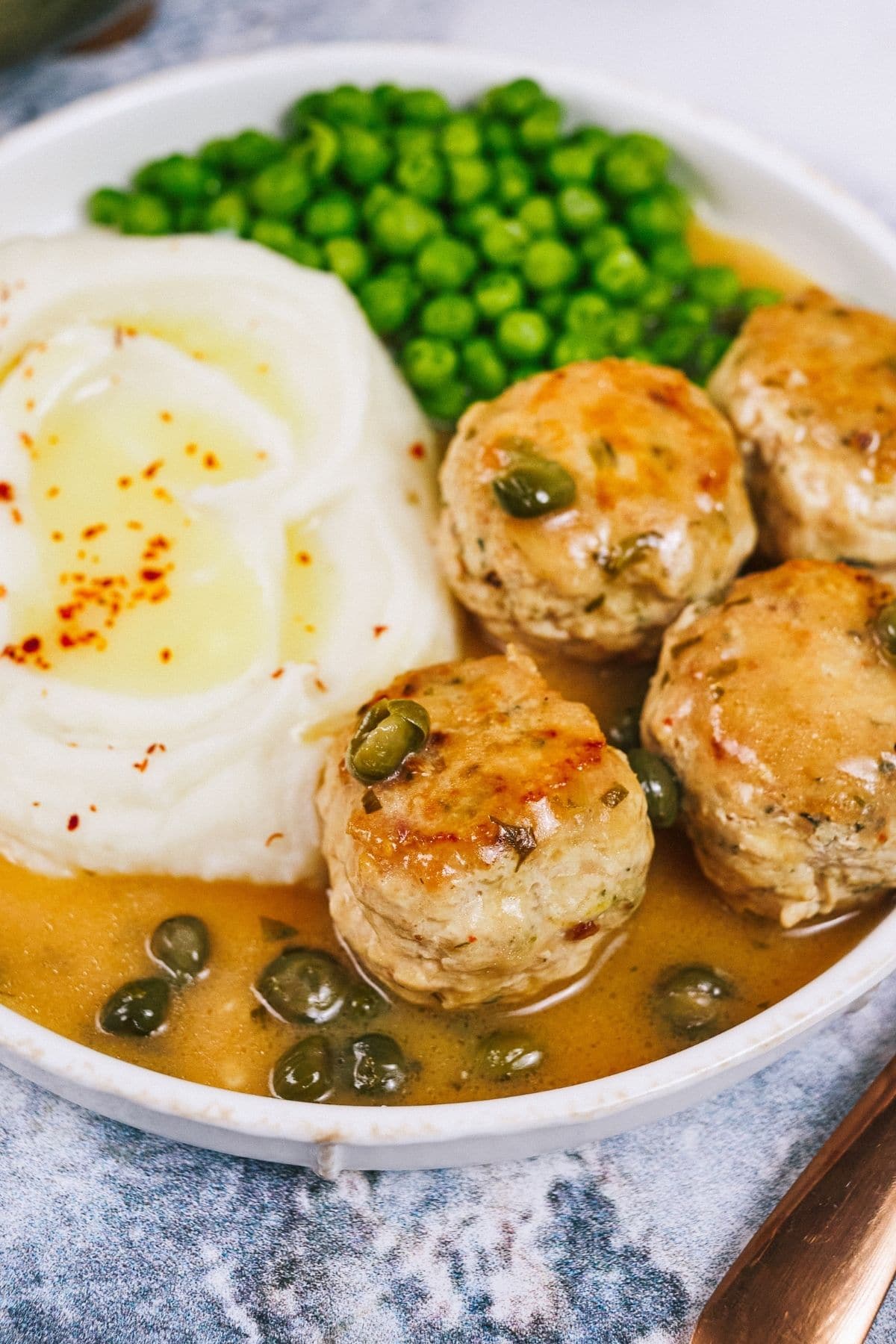 White bowl with meatballs in sauce with capers next to potatoes and peas