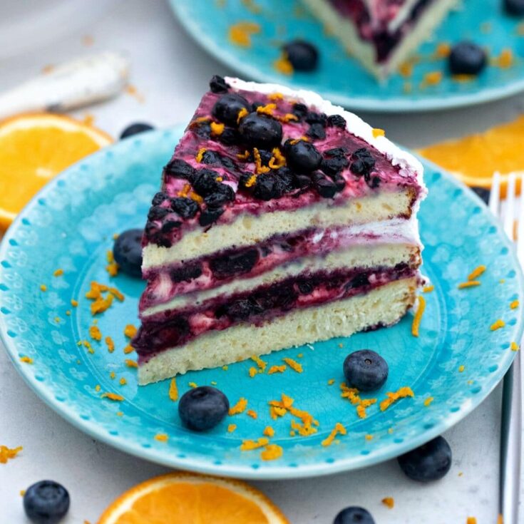 Slice of berry layer cake on light teal plate with blueberries and orange zest