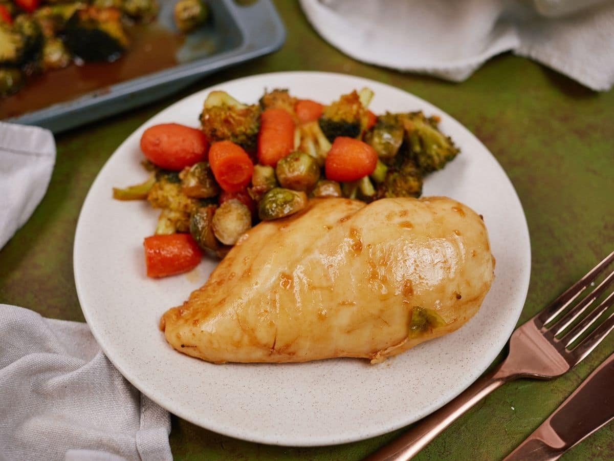 White plate with chicken breast and vegetables on green tablecloth by sheet pan