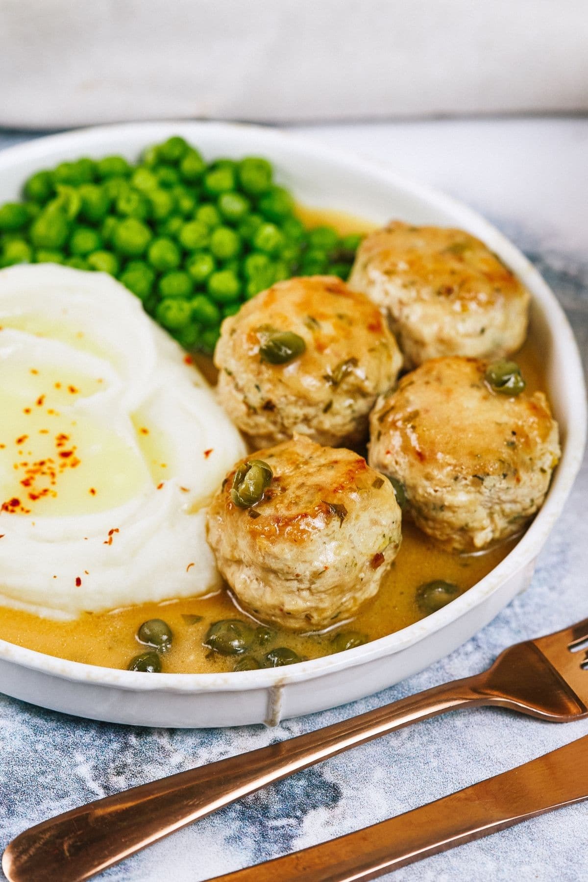 Shallow white bowl of chicken piccata metablls in sauce with mashed potatoes and peas