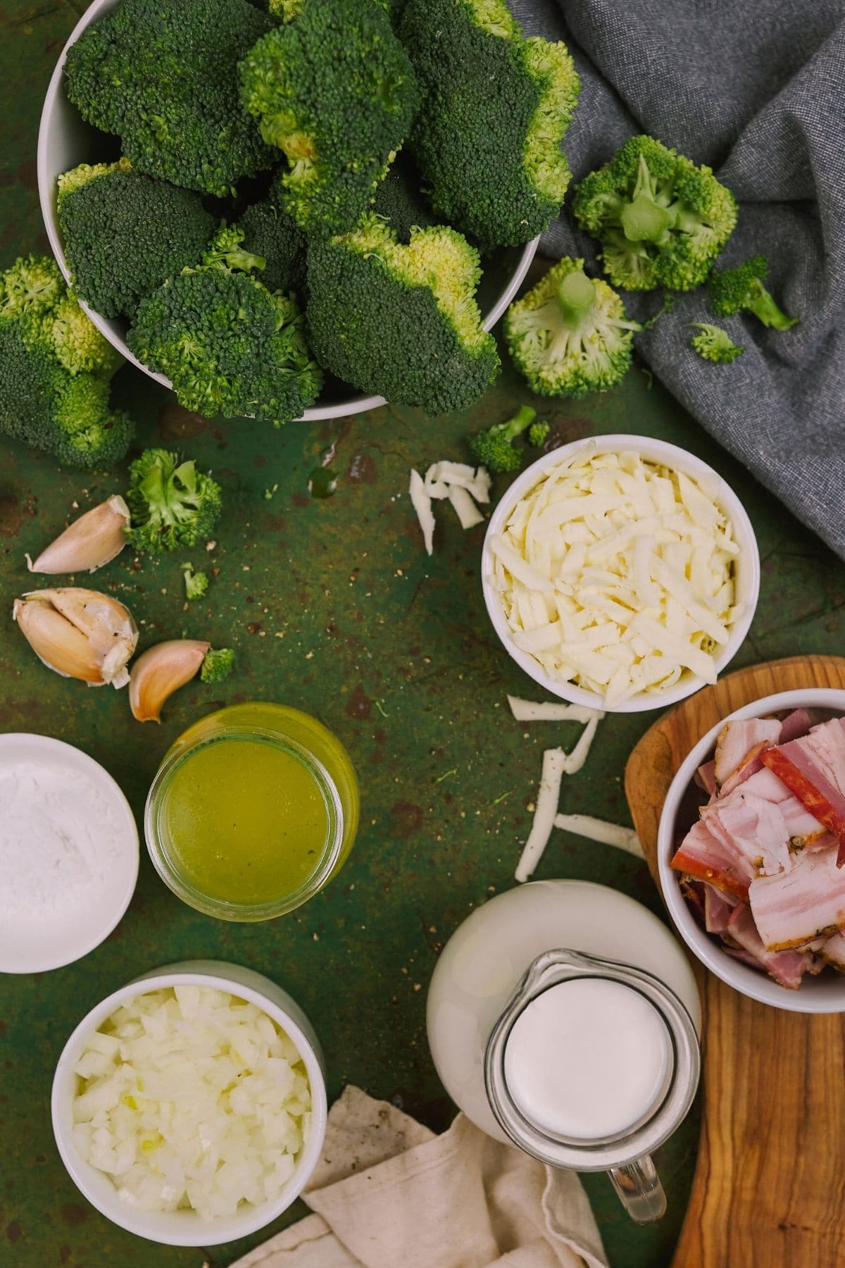 Large bowl of broccoli on green tablecloth next to bowls of cheese cream and bacon