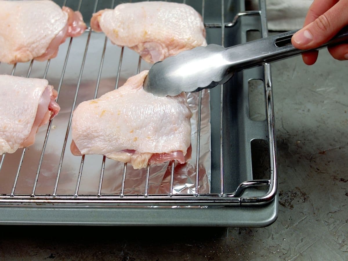 Tongs placing chicken thigh onto wire rack over baking sheet
