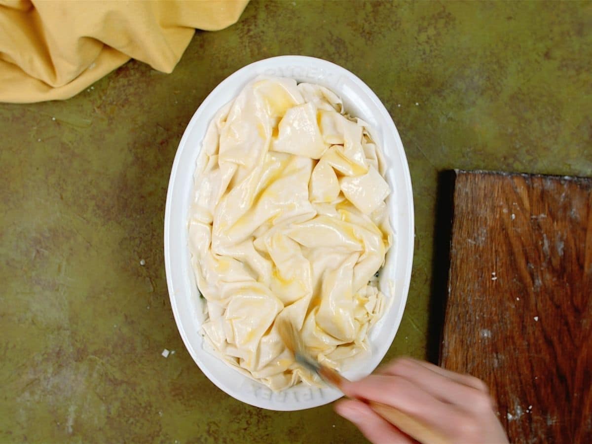 Hand brushing butter over top of phyllo dough in baking dish