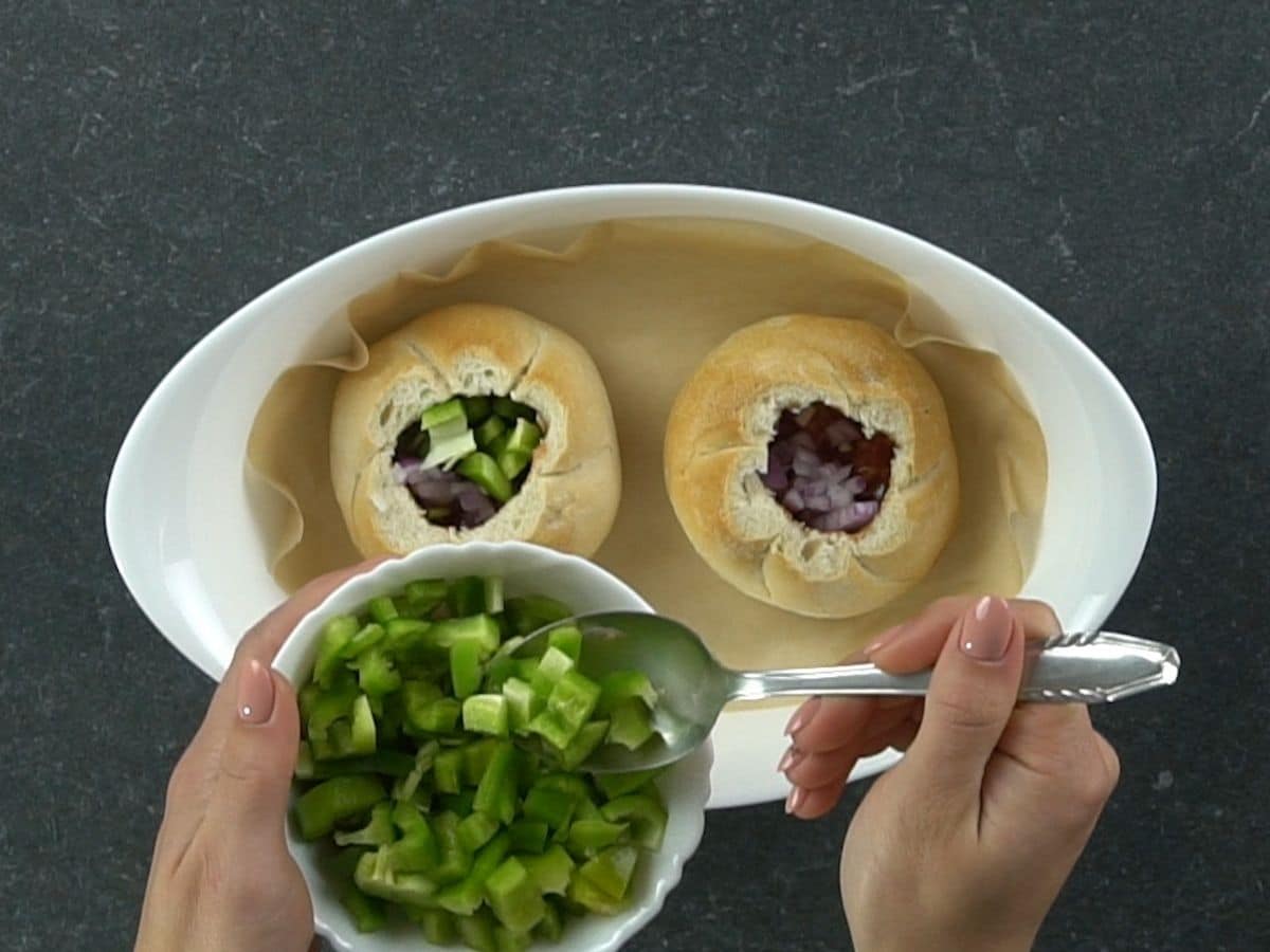 spoon of bell pepper in white bowl hovering over top of buns in white oval pan