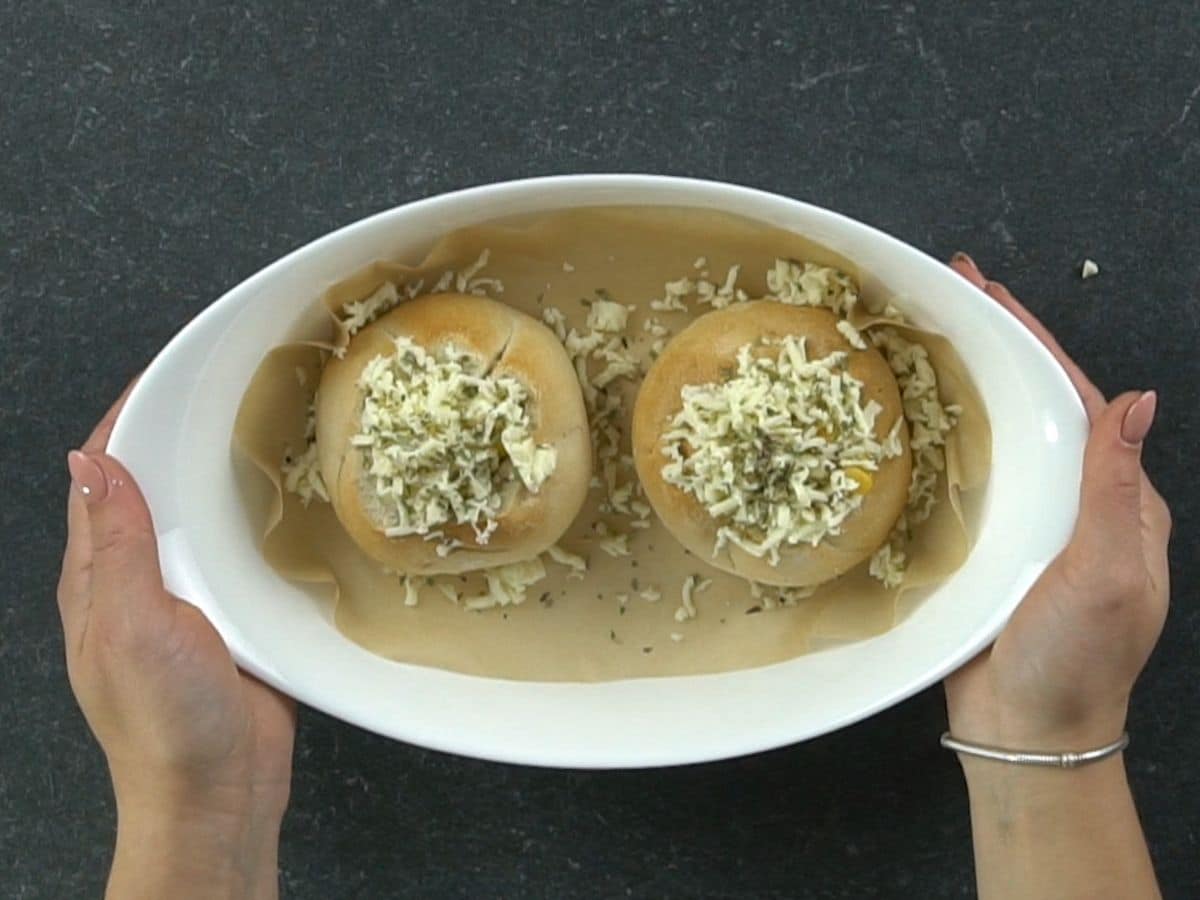 hands holding oval white baking dish with two buns inside covered in cheese