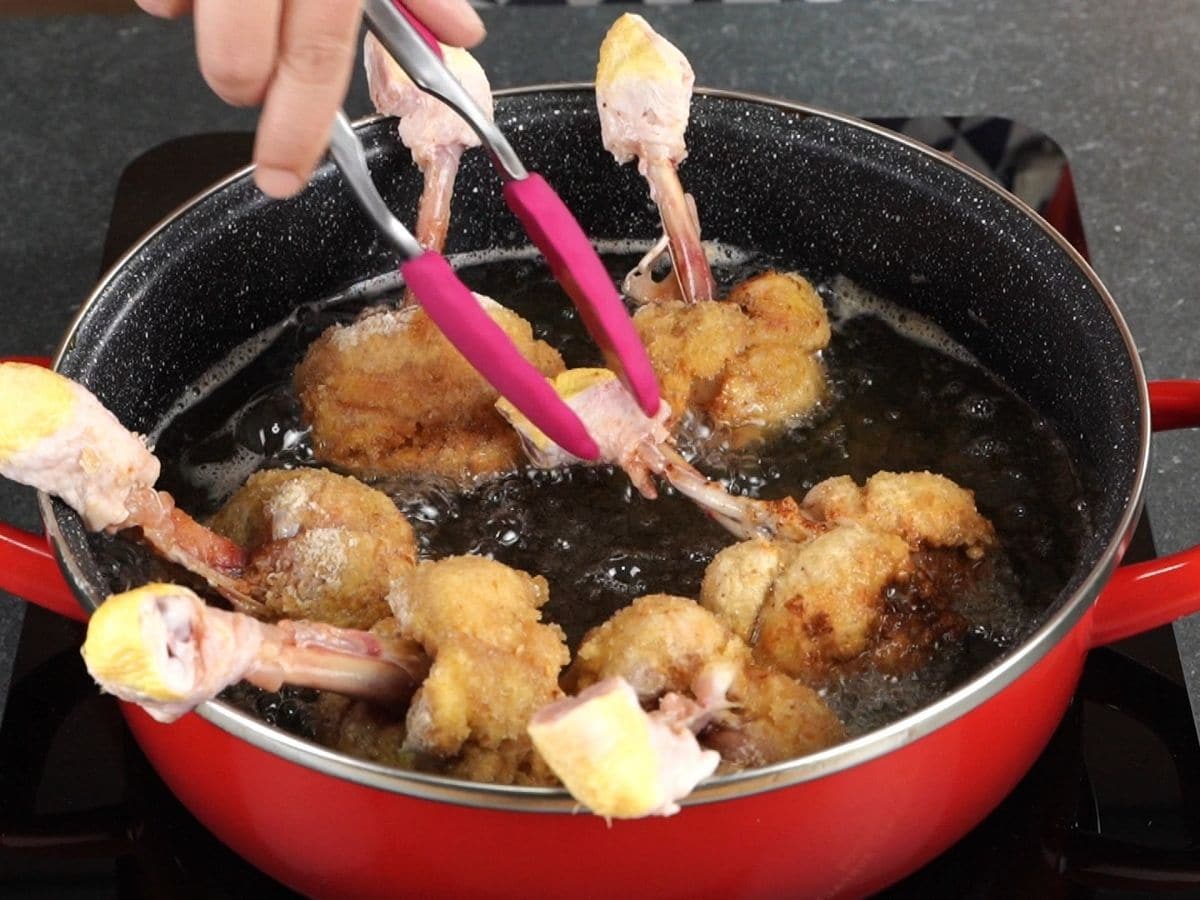 pink tongs turning chicken drumstick lollipops in hot oil in red pot
