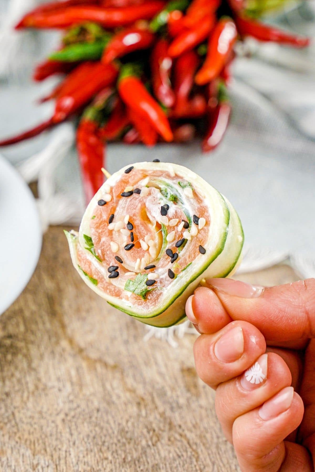 Hand holding single cucumber sushi roll by toothpick above table with red peppers in background