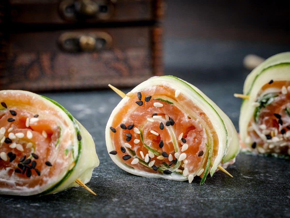 three cucumber smoked salmon rolls on black plate with sesame seeds on ends