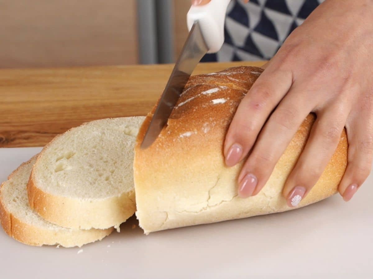 ladies hand slicing loaf of bread on white cutting board