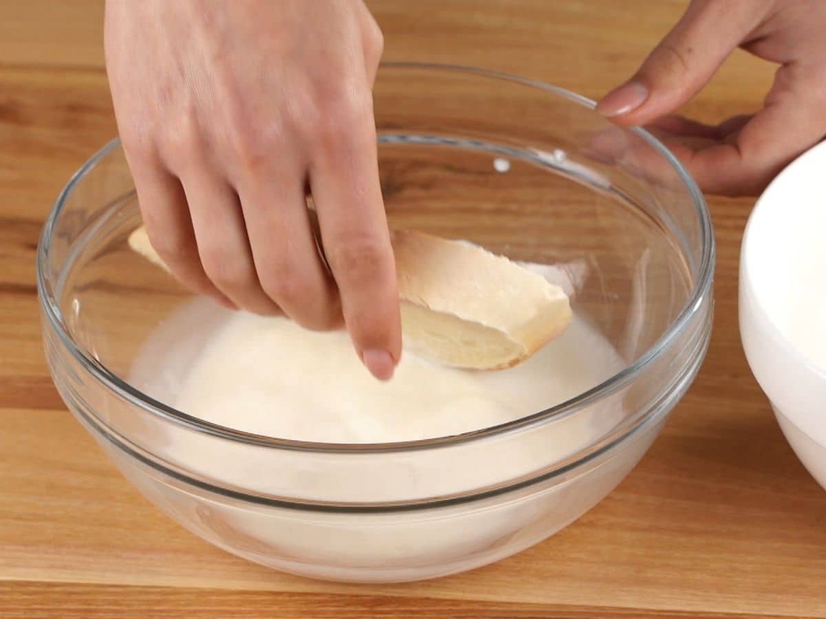 hand dipping slice of bread into milk in glass bowl