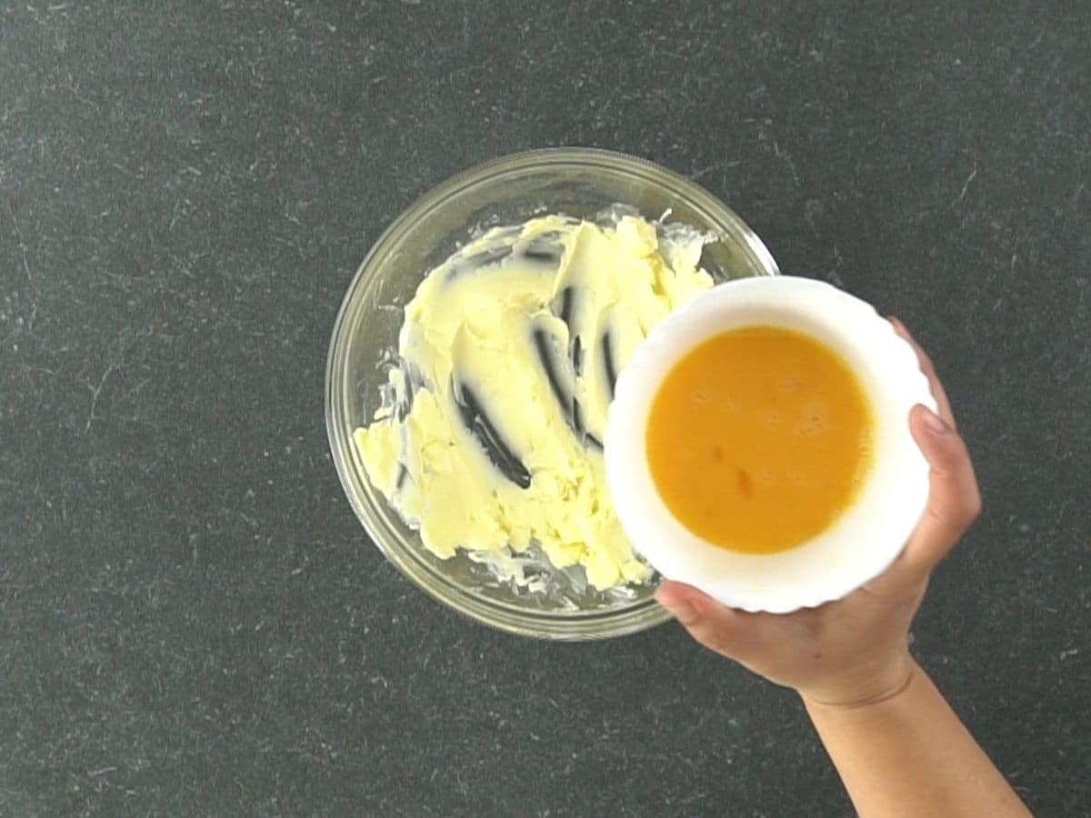 creamed butter in glass bowl with bowl of eggs being poured into butter