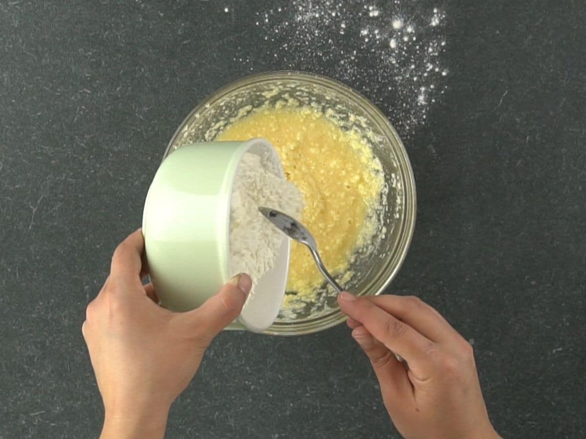hand pouring flour into glass bowl of butter and eggs