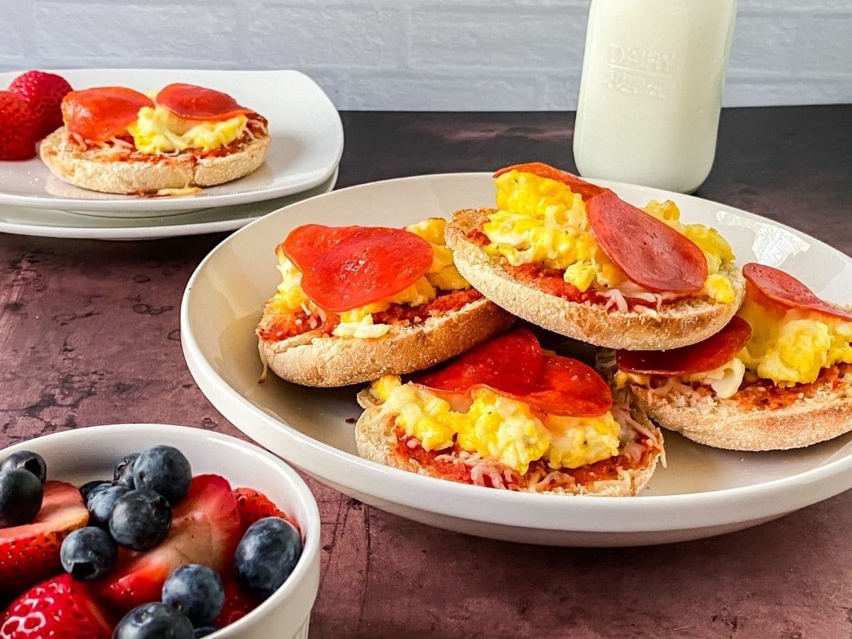 white plate of english muffin breakfast pizzas on wood table with glass of milk in background