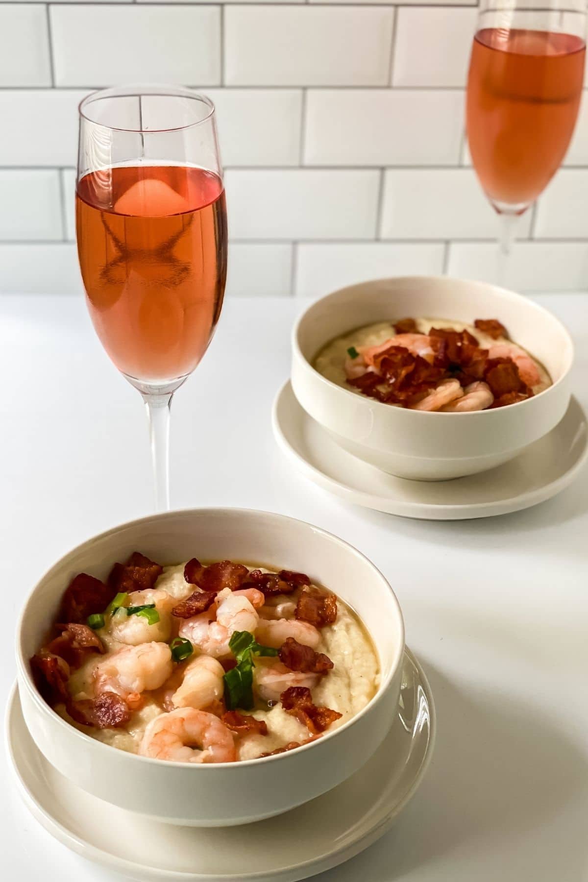 two bowls of shrimp and grits on white table with glasses of wine in background