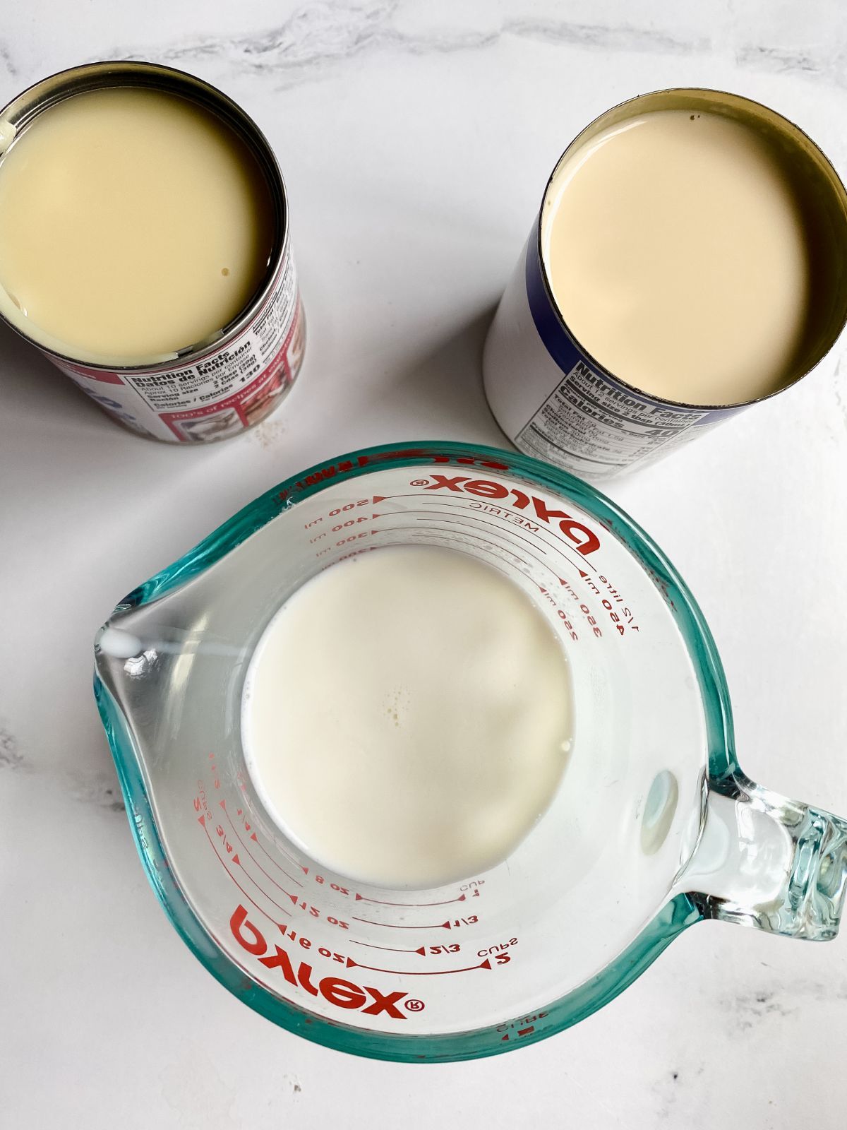 glass measuring cup with milk next to two cans of milk