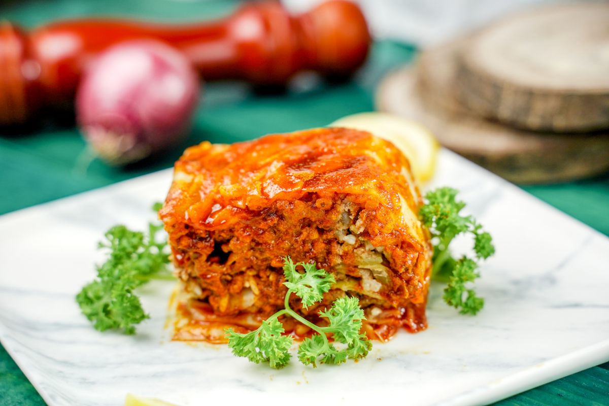 square white and gray plate with slice of lasagna on top next to red onions on teal tablecloth