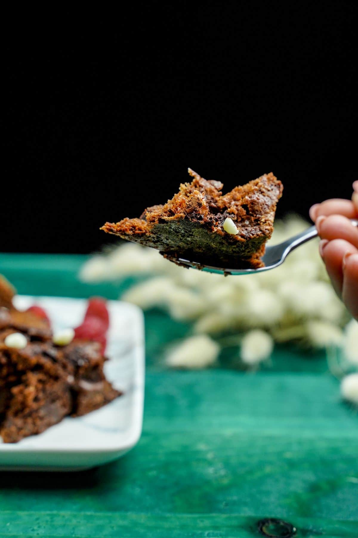 fork of brownie held above green table with plate of brownies in background