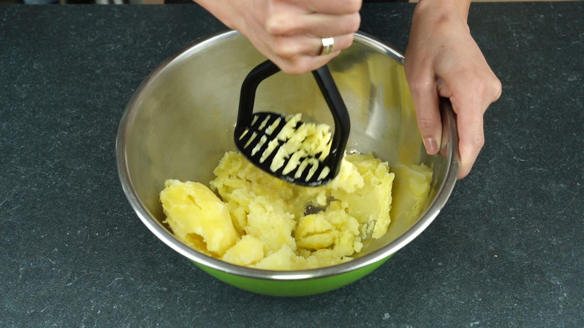 hand holding black potato masher in bowl with potatoes