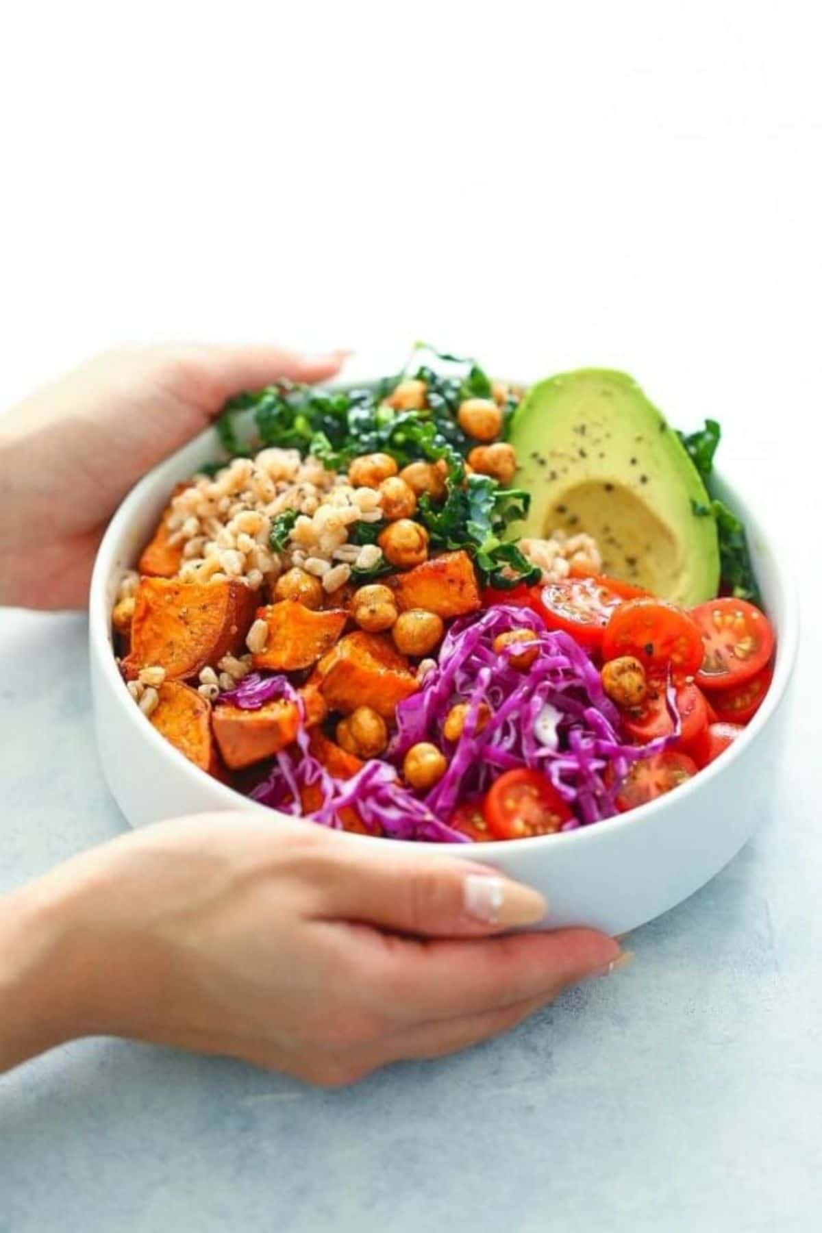 hands holding white bowl of sweet potatoes avocado and greens