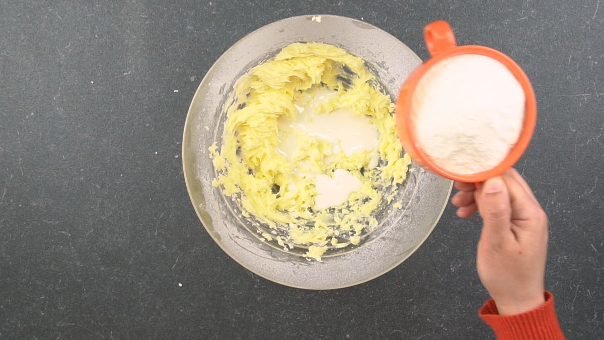 orange measuring cup of flour held above bowl of butter