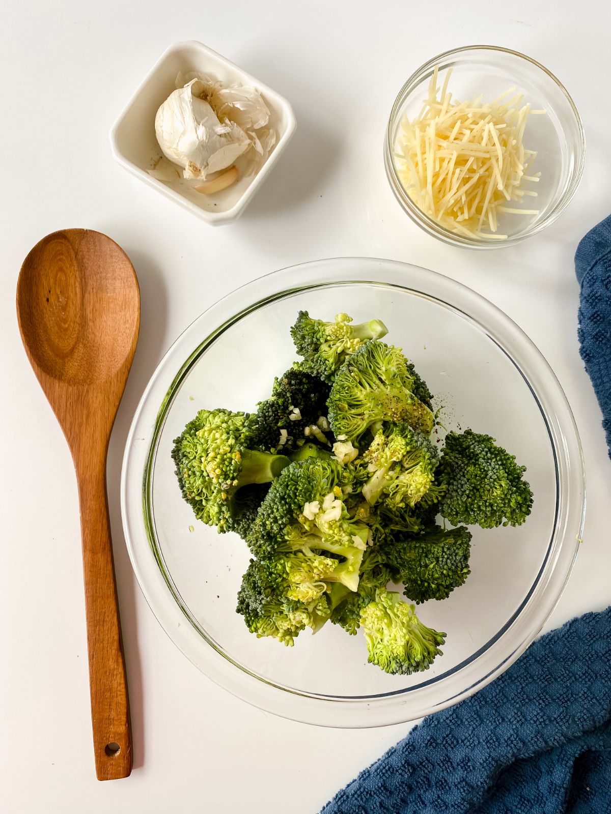 glass bowl of broccoli on white table with wooden spoon and small bowl of parmesn on the side