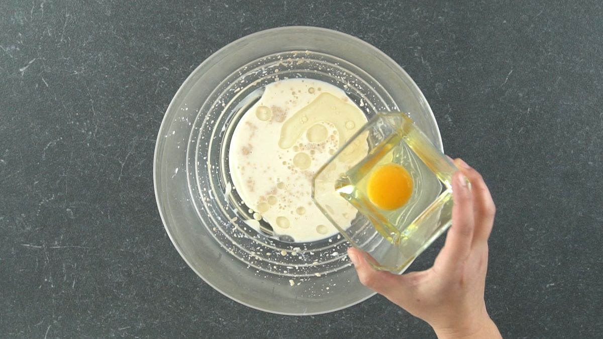 hand pouring egg into dough starter in glass bowl