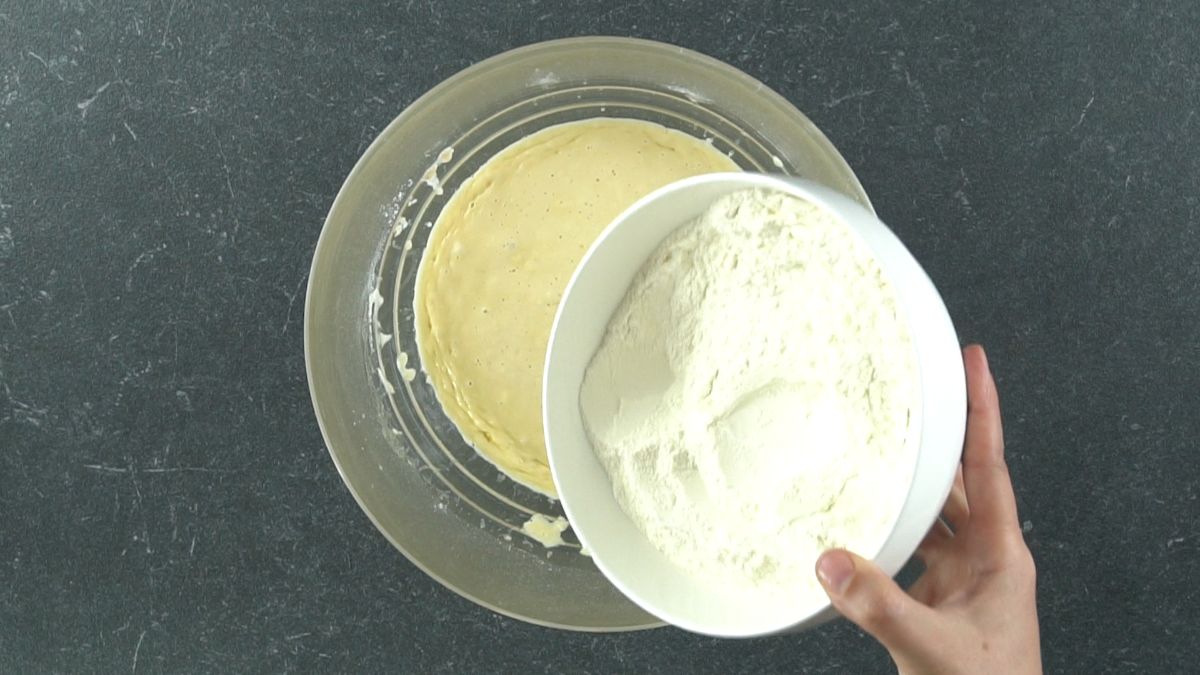 hand holding white bowl of flour above bowl of eggs and yeast