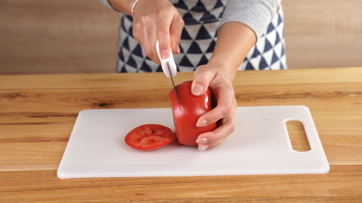 hand slicing a tomato sitting on a white cutting board