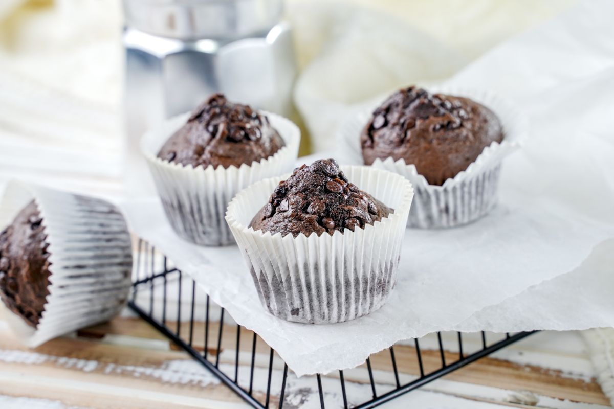 three chocolate muffins on top of black wire basket with silver coffee pot in background