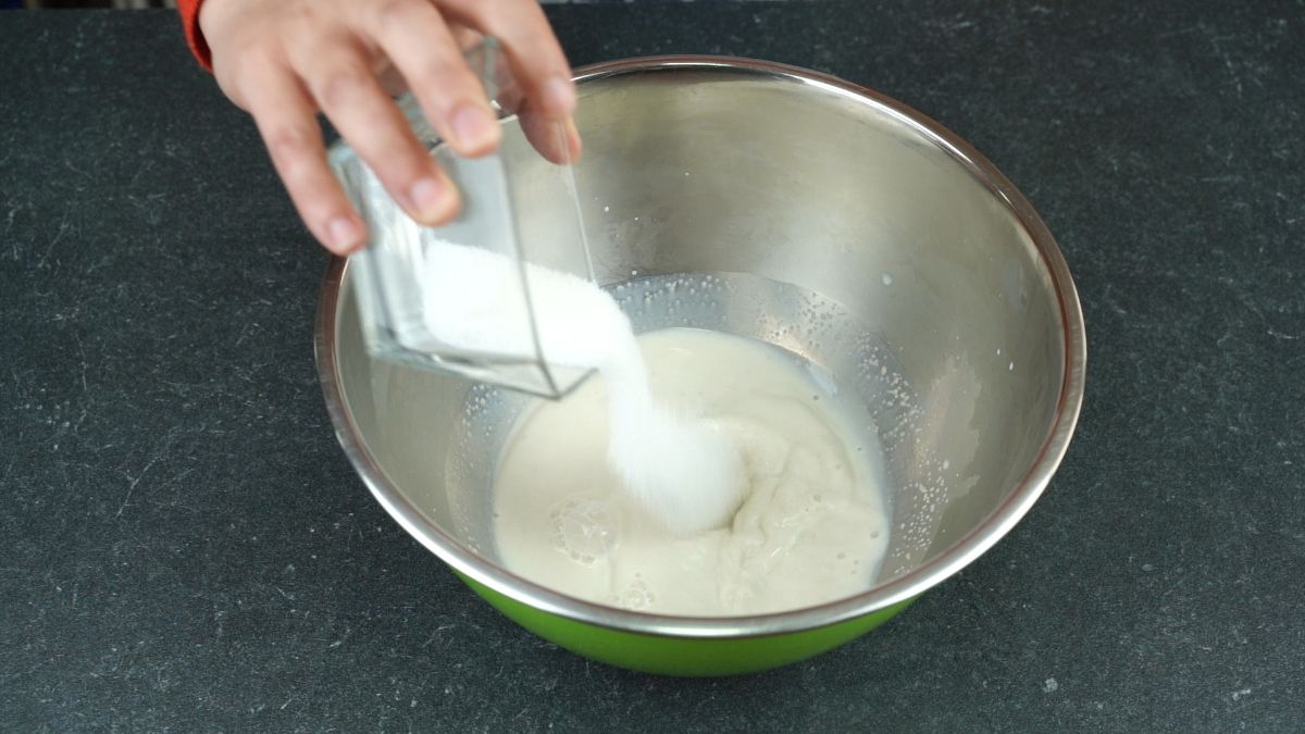 hand pouring sugar into yeast and milk in stainless steel bowl