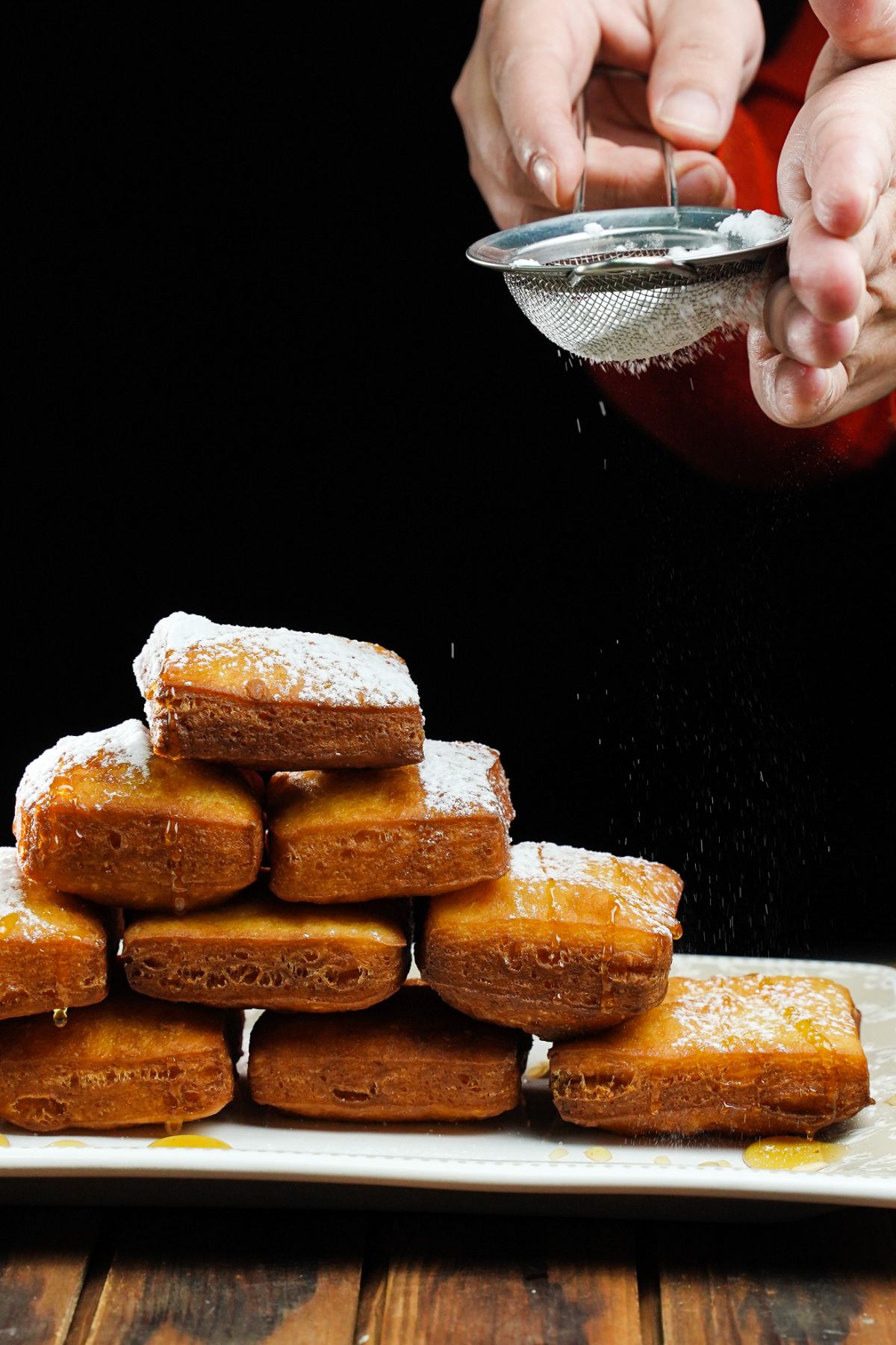tower of beignets on white platter with hand sprinkling powdered sugar over the top